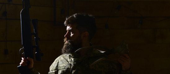 Hunter concept. Macho on strict face at gamekeepers house ready for hunting. Hunter brutal with gun, lighted in darkness, copy space. Man with beard wears camouflage clothing, dark background.