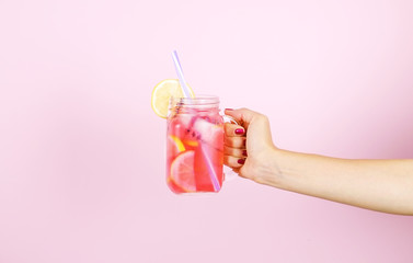 Young woman hands holding freshly squeezed strawberry lemonade of citrus fruits. Female with mason jar full of cold cocktail, lemon, orange, lime & mint leaves. Pink background, copy space, close up. - obrazy, fototapety, plakaty