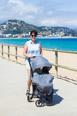 parenting and travel concept - young mother pushing baby stroller on the beach