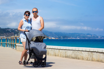 parenting and travel concept - young parents with baby stroller on the beach
