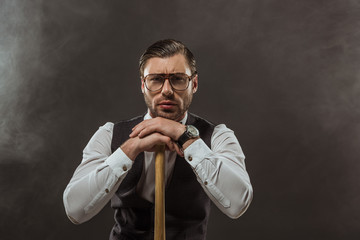 serious stylish man in eyeglasses leaning at baseball bat and looking at camera on black