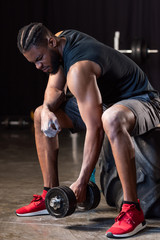 athletic african american man sitting on tyre and exercising with dumbbell