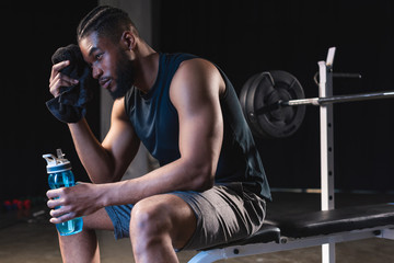 tired african american sportsman holding towel and bottle of water while sitting in gym