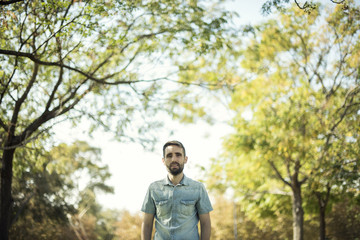 Minimalist portrait of white young man with blue shirt in a natural environment with trees, branches with leaves and a clear blue sky. Natural bucolic image.