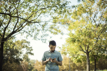 Minimalist portrait of young white man with blue shirt checking mobile in a natural environment with trees, branches with leaves and a clear blue sky. Natural bucolic image.
