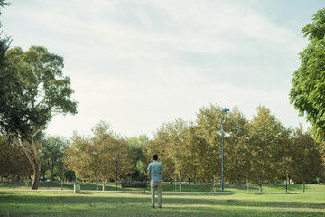 Minimalist portrait of man with his back to the distance in the middle of a landscape of a park or forest with many green trees and a large expanse of grass.
