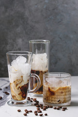 Iced coffee cocktail or frappe with ice cubes and cream served in three different glasses with coffee beans around on white marble table with grey concrete wall at background.