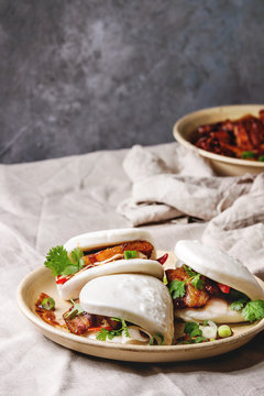 Asian sandwich steamed gua bao buns with pork belly, greens and vegetables served in ceramic plate on table with linen tablecloth. Asian style fast food dinner.