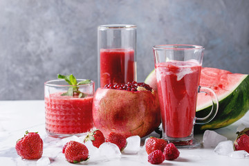 Three different red fruit berry watermelon, strawberry, raspberry, pomegranate cocktails or smoothies in glasses with crushed ice, fresh mint and ingredients above on white marble table.