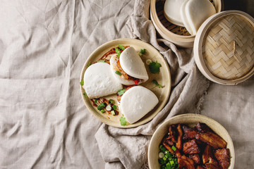 Asian sandwich steamed gua bao buns with pork belly, greens and vegetables served in ceramic plate over linen tablecloth. Asian style fast food dinner. Flat lay, space