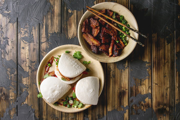 Asian sandwich steamed gua bao buns with pork belly, greens and vegetables served in ceramic plate over dark wooden plank background. Asian style fast food dinner. Flat lay, space