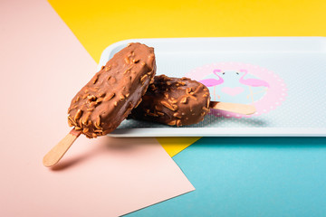 Dessert chocolate ice cream bars with nuts, wooden stick on blue and yellow pastel background. Eskimo in blue vintage tray with pink flamingosand dots. Summer temptation. Indoors closeup.