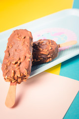 Dessert chocolate ice cream bars with nuts, wooden stick on blue and yellow pastel background. Eskimo in blue vintage tray with pink flamingosand dots. Summer temptation. Indoors closeup.