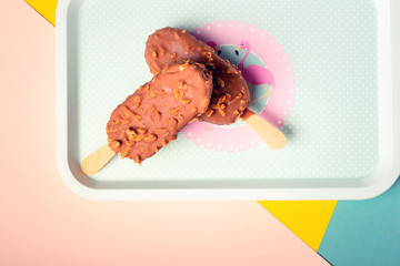 Dessert chocolate ice cream bars with nuts, wooden stick on blue and yellow pastel background. Eskimo in blue vintage tray with pink flamingosand dots. Summer temptation. Indoors closeup.