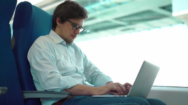 Young manager wearing a white shirt and jeans is riding a train to work and using his laptop. Handheld real time medium shot