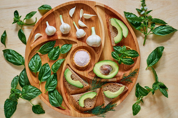 Avocado toasts on the wooden plate with  garlic, basil, dill and salt. Vegan avocado sandwiches decorated with basil leaves. Ingredients for healthy breakfast or summer green diet. Top view