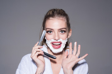 Girl on smiling face wears bathrobe, grey background. Lady play with sharp blade of straight razor. Woman with face covered with foam holds straight razor in hand. Barber and shaving concept