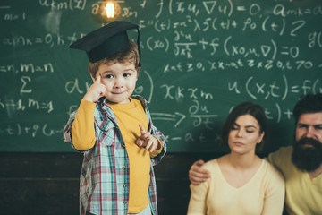 Smart child, wunderkind in graduate cap pointing at his head. Boy presenting his knowledge to mom and dad. Parents listening their son, chalkboard on background. Wunderkind concept