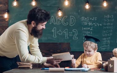 Wunderkind and genius concept. Father, teacher reading book, teaching kid, son, chalkboard on background. Boy child in graduate cap likes to listening dad. Dad wants to grow up genius son