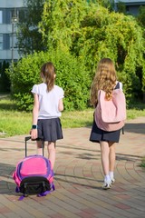Rear view of two schoolgirl girlfriends elementary school students walking with school bag in the yard