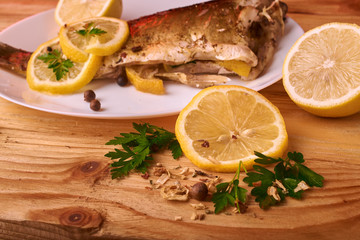 Baked fish with lemon, spices and herbs on white plate on wooden background without head