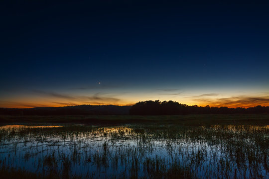 A bright sunset in the night sky. The landscape with the river and trees is photographed on a long exposure.