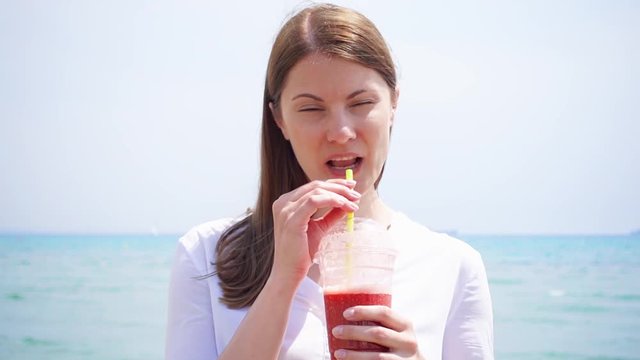 Smiling vegan woman in white shirt drinking from cup with strawberry smoothie against sea in slow motion. Fit vegetarian female enjoying healthy lifestyle outdoors