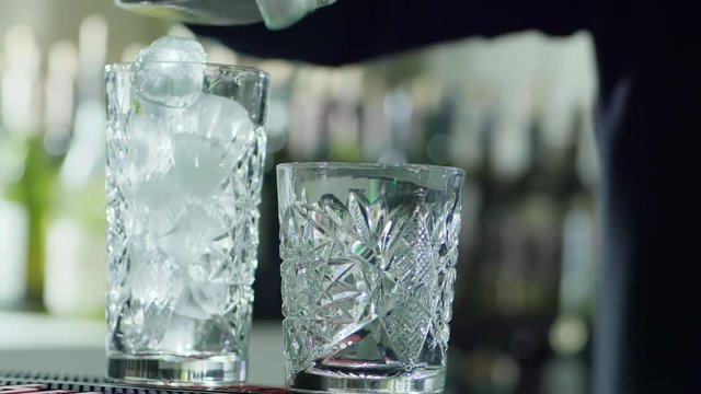 bar worker holds metal scoop in arms and strew ice cubes into transparent glass close-up on desk on unfocused background