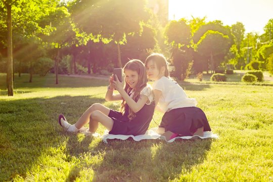 Cheerful little girl friends play with smartphone sitting on a meadow in the park. The concept of people, children, technology, friends and friendship