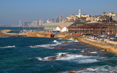 Zatoka Morza Śródziemnego w Tel Awiwie-Jaffie, promenada nadmorska w starej Jaffie, budynki, zaparkowane samochody, skały w wodzie, w tle, nieco rozmyte, drapacze chmur nowoczesnej częsci miasta - obrazy, fototapety, plakaty