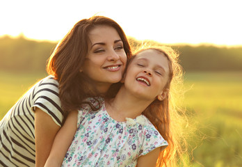 Emotional calm mother hugging her laughing kid girl with loving eyes on sunset bright summer background. Closeup bright color portrait