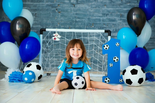 Little cheerful child dressed in sports clothes sitting on the floor near a football goal, looking at a big soccer ball. The first year, number one