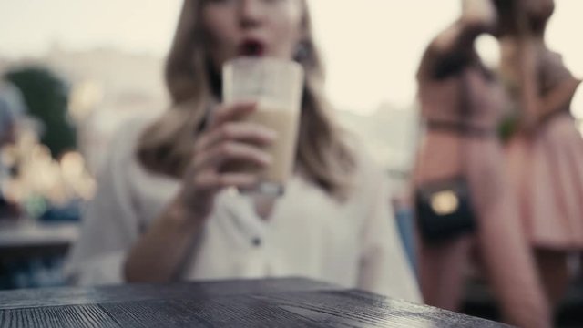 In the defocus, a young Caucasian blonde woman, curly hair, with sexy red lips drinks an alcoholic cocktail through a black straw from a large bar glass in a restaurant on a city street. Lonely