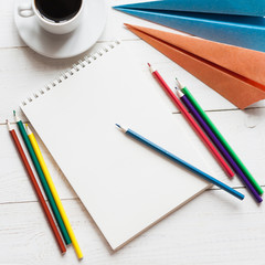 A blank sheet of paper, colored pencils, paper airplanes and a cup of coffee. The concept of a dream of traveling or planning a vacation, copy space. A square photo.