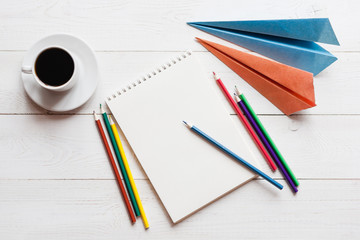 A blank sheet of paper, colored pencils, paper airplanes and a cup of coffee. The concept of a dream of traveling or planning a vacation, copy space.