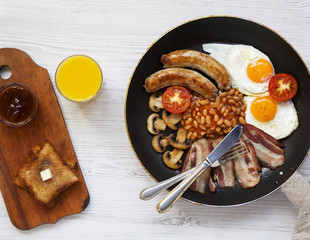 Full English breakfast in a pan with fried eggs, bacon, beans, sausages  and toasts on a white wooden background, top view. From above, overhead.