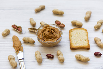 Bowl of peanut butter with peanuts and knife over white wooden background, closeup. Side view.