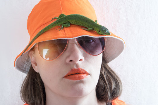 Slender girl in orange fashionable semi-overalls, orange hat and yellow household gloves, with sunglasses holding live green lizard and to playing with her. Concept care, careful attitude for animals