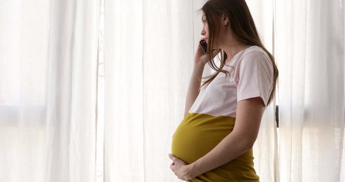 Side view of casual woman in expectancy having phone call while standing near window with white curtains