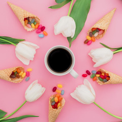 Composition of bright candy in waffle cones, cup of coffee and white tulip flowers on pink background. Flat lay, top view