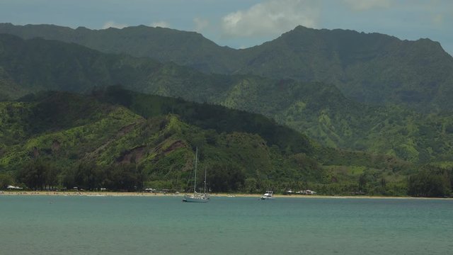 Hanalei Bay, on the Hawaiian island of Kauai, is shown in a daytime view, with boats in the foreground waters and green, lush mountains in the background.
