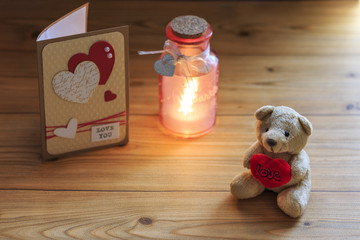 Small teddy bear with heart and bottle light and greeting card with hearts on wood table