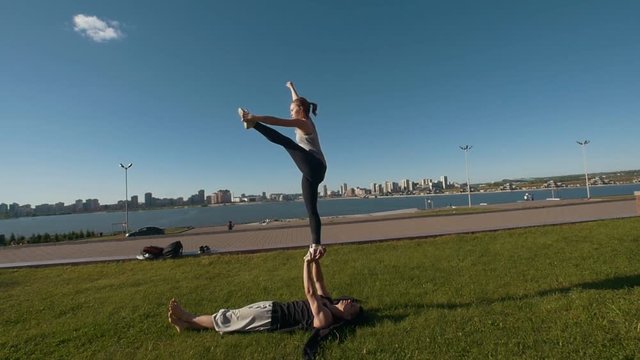 Young sporty couple practicing acrobatics performing front plank bird pose, young man holding flying woman balancing on his arms outdoors at sunset