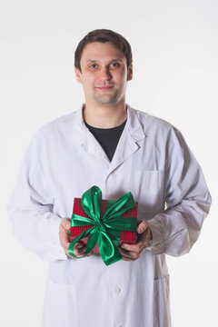 a man in a white robe holds a gift box. smiling doctor with a red box with a green bow. Handsome man looking at camera on white background