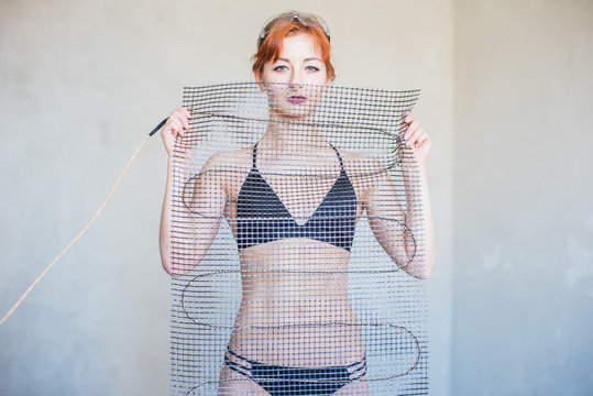 A girl with red hair dressed in a black swimsuit, holding a set for a warm floor. Thick electrical cable on the grid.