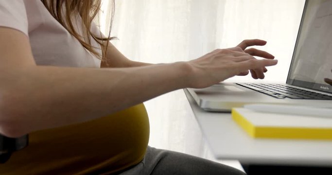 Faceless shot of pregnant woman in expectancy sitting at table with laptop and taking care of business in daylight