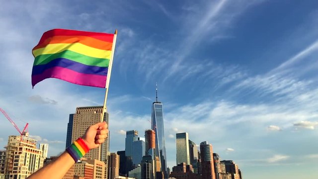 Hand wearing gay pride wrist band holding rainbow flag up in front of the city skyline at sunset