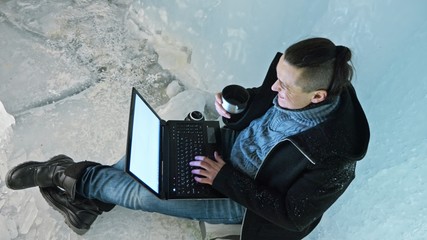 Man is sit on the internet in laptop in an ice cave. Around the mysterious beautiful ice grotto. User communicates in social networks. He crazy strange hipster in black coat and fashionable hairdo - Powered by Adobe