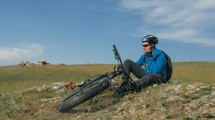 Naklejka na ściany i meble Fat bike also called fatbike or fat-tire bike in summer driving through the hills. The guy sits on a stony sandy and grassy hill after a trip. He rests and enjoys the fresh air.