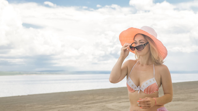Attractive woman in a hat stands on the beach against the blue sea. She has sunglasses and a panama on her head. On her body is a swimsuit and a silk cape. In the background are very beautiful deep
